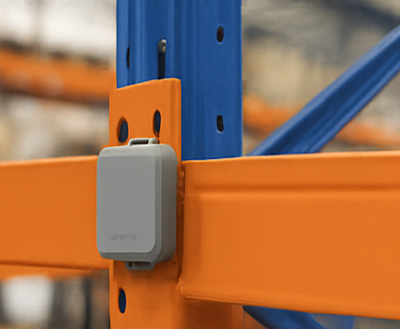 A Square Impact sensor screwed to the upright of an orange and blue pallet rack, level with a beam, in a warehouse aisle